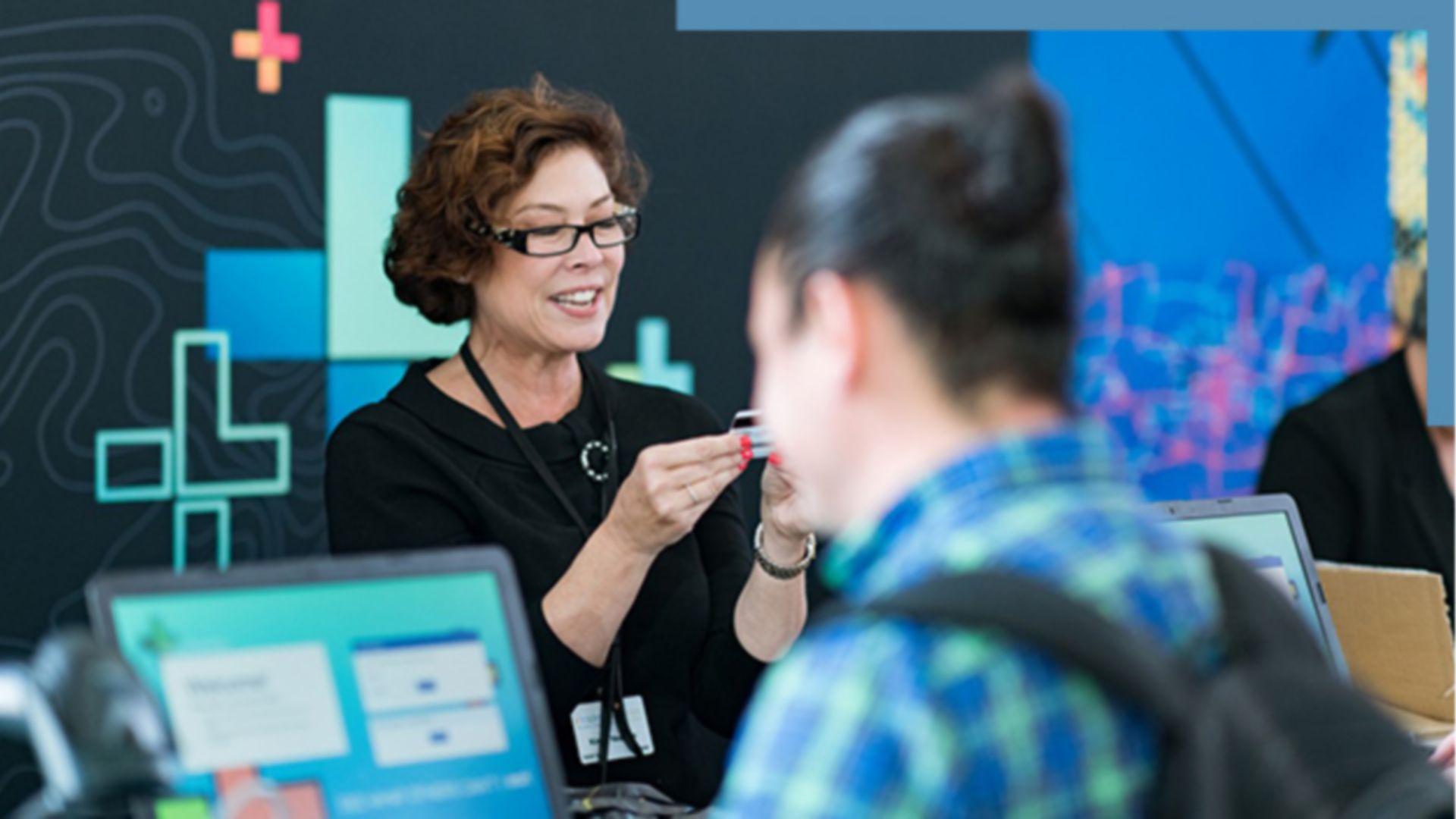 A photo of a conference staffer helping a registrant, while a person in a backpack stands in the foreground using a laptop