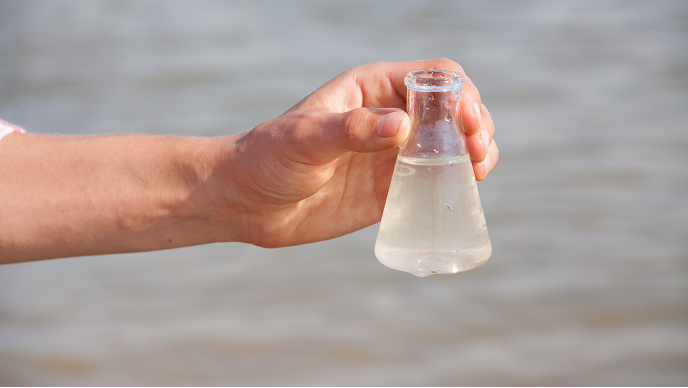 A hand holding a glass beaker partially filled with cloudy colorless fluid