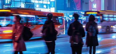 Four casually-dressed pedestrians watching brightly lit buses speed past on a busy city street at night
