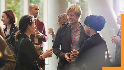 Groups of people standing together, holding beverages, and talking to each other in a sunlight-filled indoor area