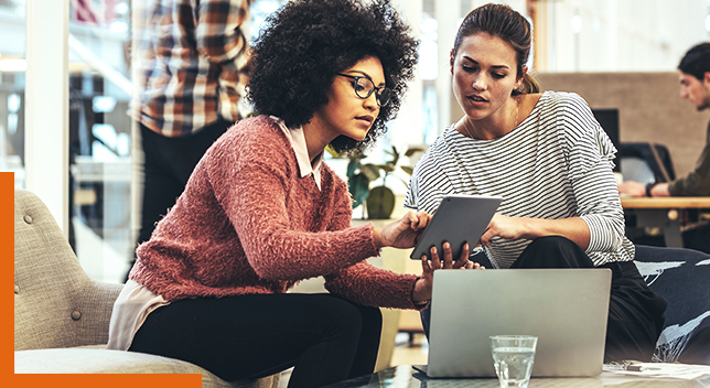 Two casually-dressed people sitting in an sunlit office lounge space sharing a tablet and discussing its display