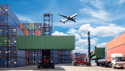A shipping hard with blue storage crates stacked high as an airplane flies low overhead through a cloud-swept blue sky