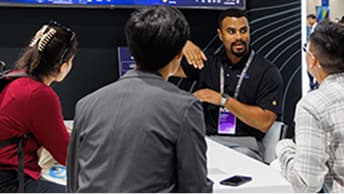 Group of three attendees seated at a table listening to a presenter in front of a dark display with graphics and text