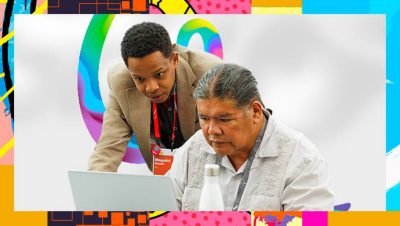 Esri expert wearing a tan suit looking over the shoulder of a seated attendee working on a laptop