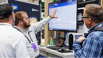 Attendee gestures toward a large monitor displaying software while two others watch