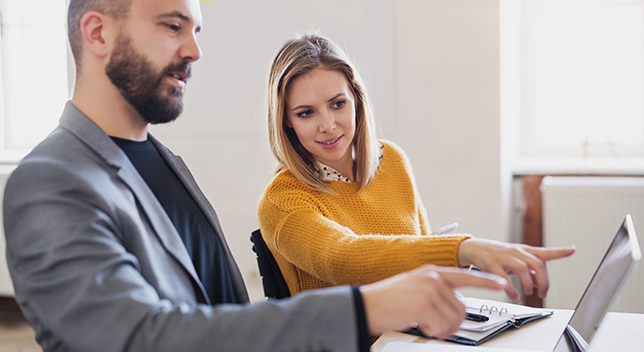 Dwóch biznesmenów siedzi w biurze i omawia niewidoczne dla czytelnika elementy na ekranie laptopa.