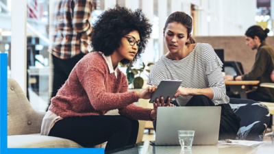 Two people looking at a tablet  one of them is holding with an open laptop on a low table in front of them
